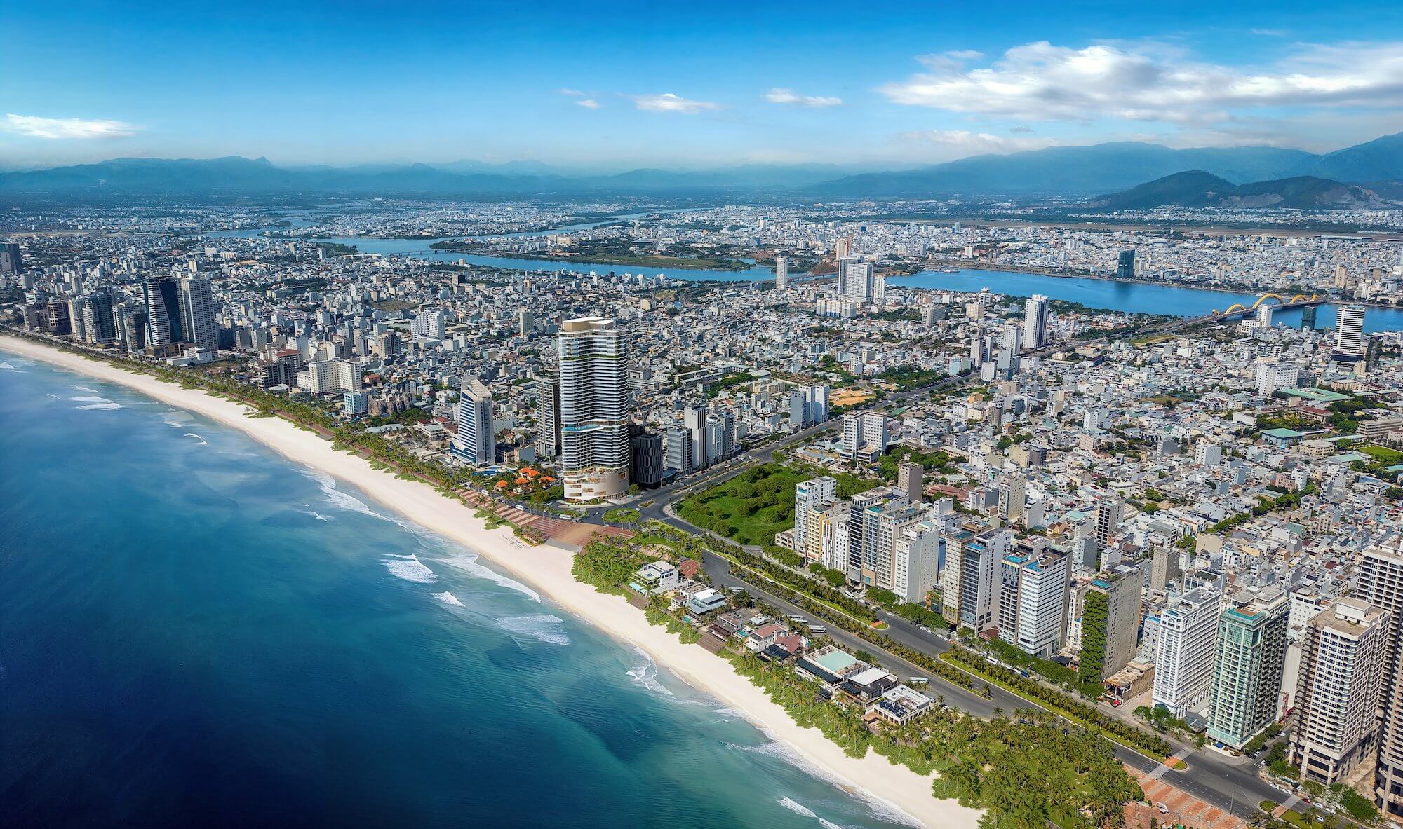 Aerial view of Nobu Residences Danang on beachfront Mỹ Khê — the tower, Danang city, the Han River, the Dragon Bridge and the mountains
